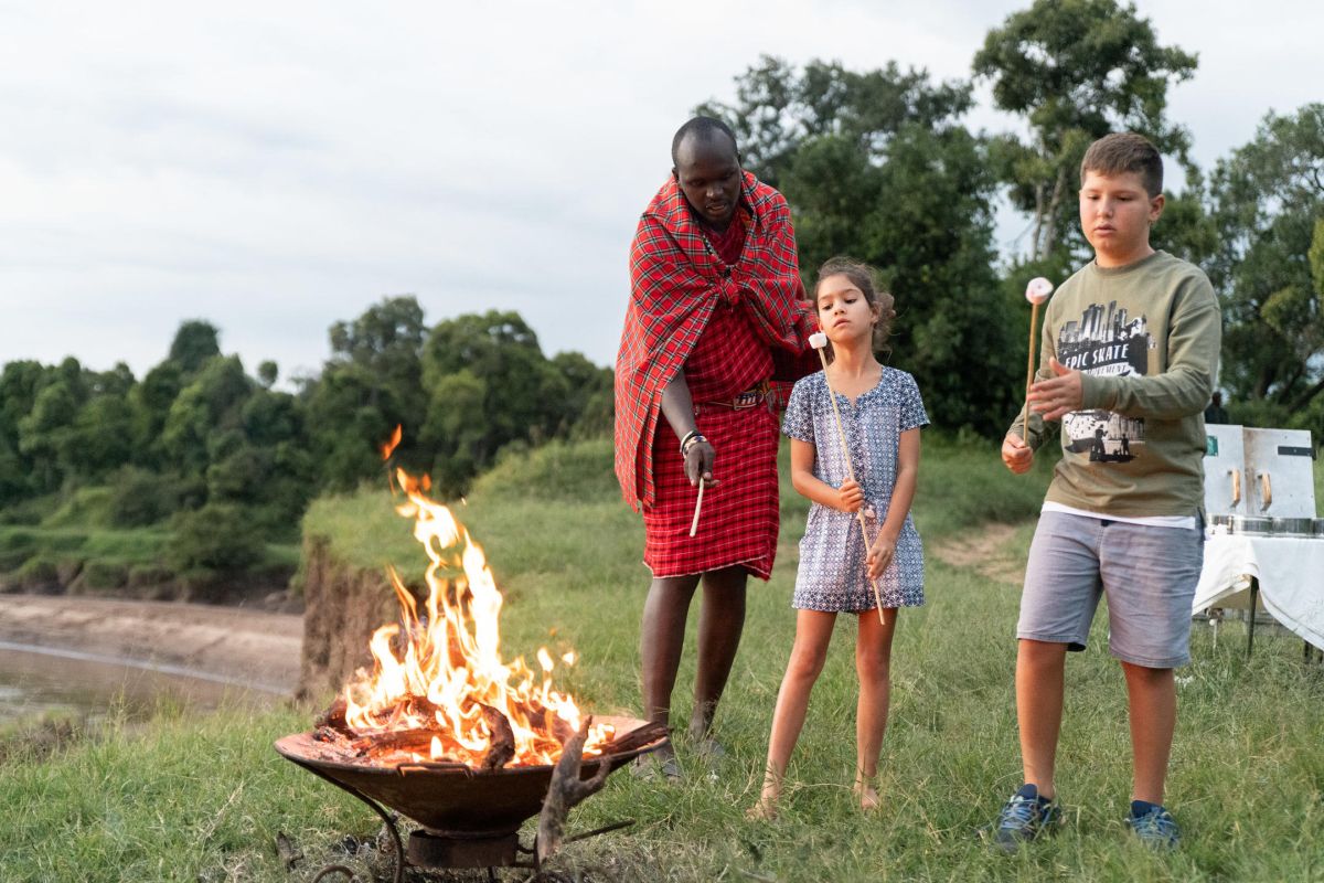 Kinder und ein Safari-Guide stehen am Lagerfeuer im Governors Camp mit Blick auf die Natur.