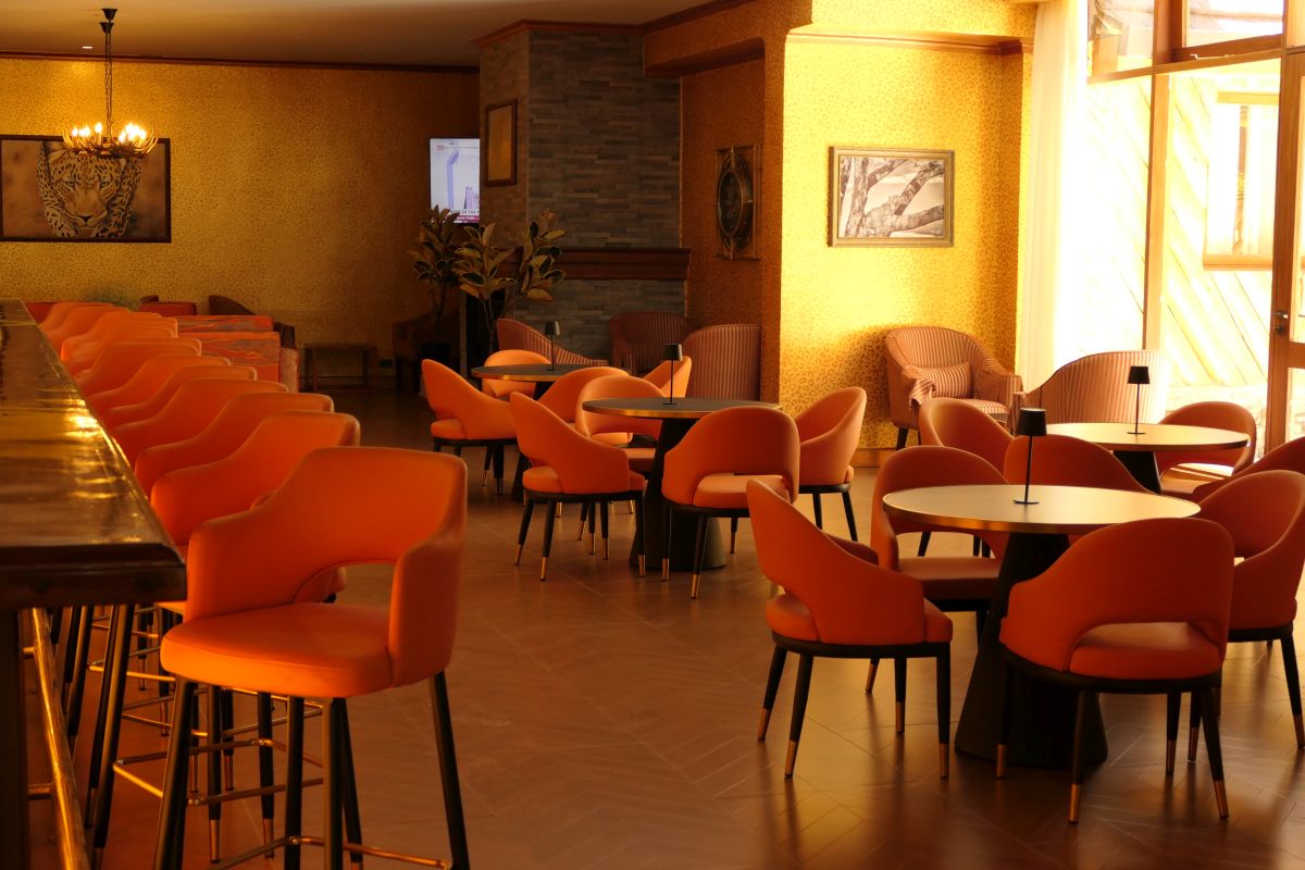 Stilvoller Bar- und Loungebereich der Sawela Lodge mit orangefarbenen Sesseln, hohen Barhockern, kleinen Tischen und großen Fenstern, die viel Tageslicht hereinlassen.